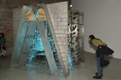 A large metal gate like sculpture with blue neon lights sits in a corner of a room. A person bends down to get a closer look. 
